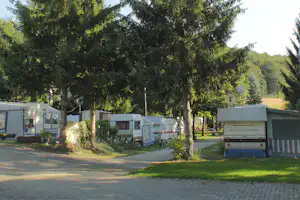 Wohnwagen auf einem Campingplatz zwischen hohen Bäumen, mit gepflastertem Weg und weiteren Stellplätzen im Hintergrund.