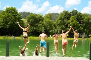 Familie springt vom Steg ins grüne Seeufer, umgeben von Bäumen und einem Spielbereich am Campingplatz.