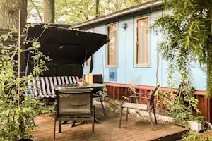 Wohnwagen auf Holzterrasse mit Grill und Sitzbank, umgeben von Bäumen und Pflanzen im grünen Campingbereich.