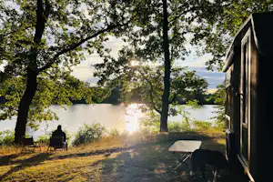 Campingwagen am See im Wald, Liegestuhl und Tisch am Weg, Bäume im Vordergrund, ruhiges Wasser im Hintergrund.