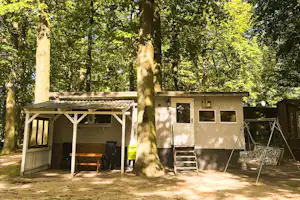 Wohnwagen auf einem Campingplatz im Schatten hoher Bäume, mit überdachter Terrasse, Tisch und Stühlen.