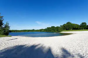 Blick auf den See mit Sandstrand am Ufer, umgeben von Bäumen und grünem Gras unter klarem Himmel.