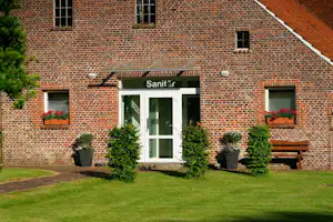 Sanitärgebäude aus Ziegelmauerwerk mit Eingangstür, Fenstern und Blumenampeln, davor Rasen und Bank am Weg.