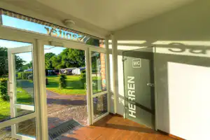 Eingang zur WC-Anlage am Campingplatz, Blick von der Terrasse auf den gepflasterten Weg und den Rasen mit Bäumen.