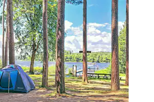 Zelt auf einem Campingplatz am See, umgeben von hohen Bäumen, mit Blick auf Steg und Wasser.