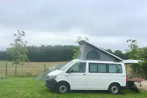 Weißer Camping-Van mit Aufstelldach und Seitenzelt auf einer Wiese, im Hintergrund Bäume und ein Zaun.