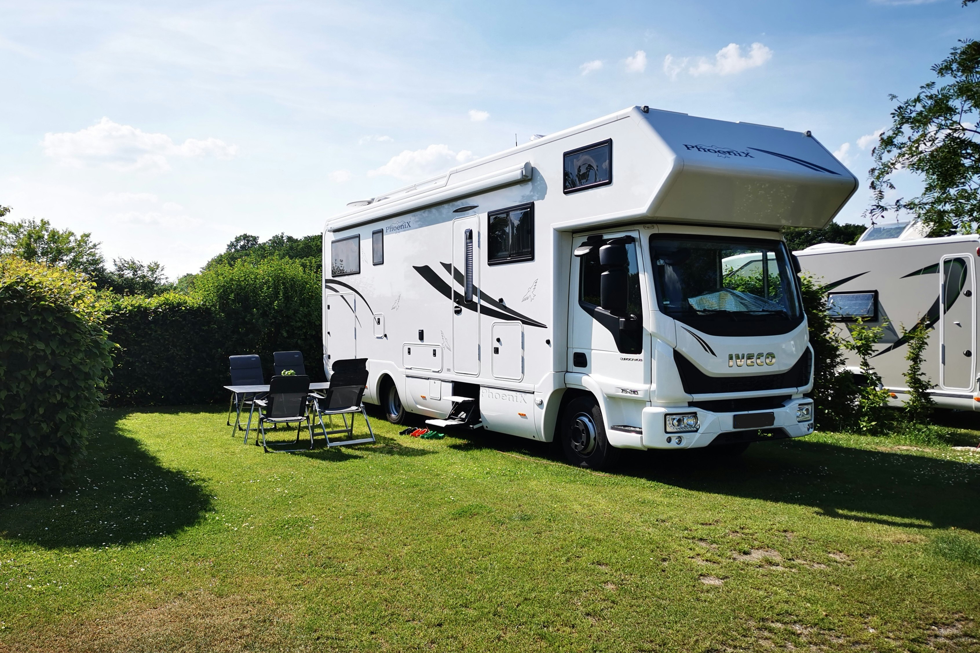 Wohnmobil auf einem grünen Campingplatz mit Markise, Sitzgruppe und Hecke im Hintergrund.