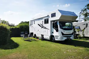 Wohnmobil auf einem grünen Campingplatz mit Markise, Sitzgruppe und Hecke im Hintergrund.