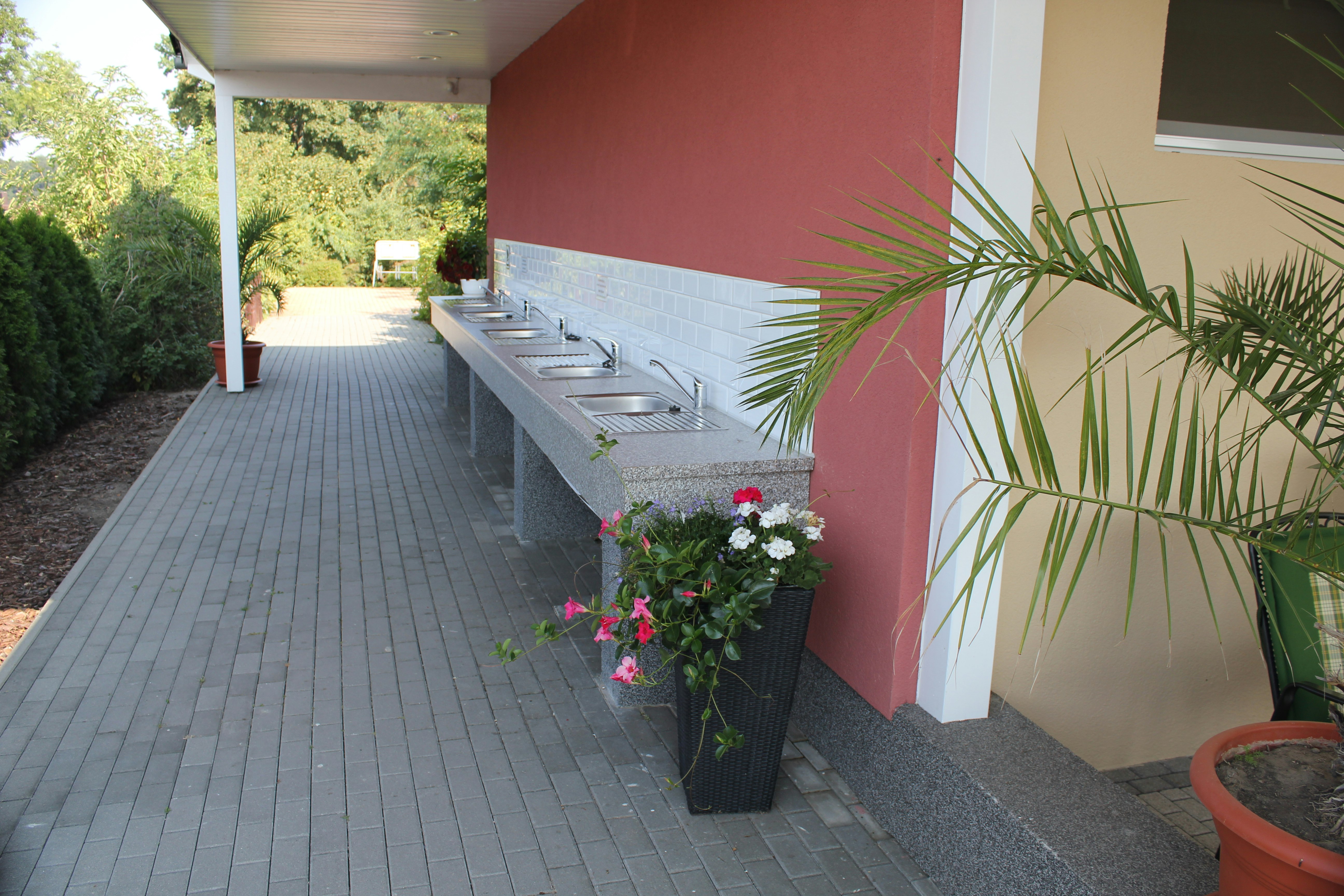 Überdachter Gehweg am Campingplatz neben einer Küchenzeile mit Spüle, Blumen und Pflanzen vor einem roten Gebäude.