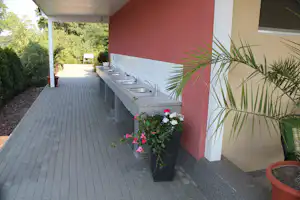Überdachter Gehweg am Campingplatz neben einer Küchenzeile mit Spüle, Blumen und Pflanzen vor einem roten Gebäude.