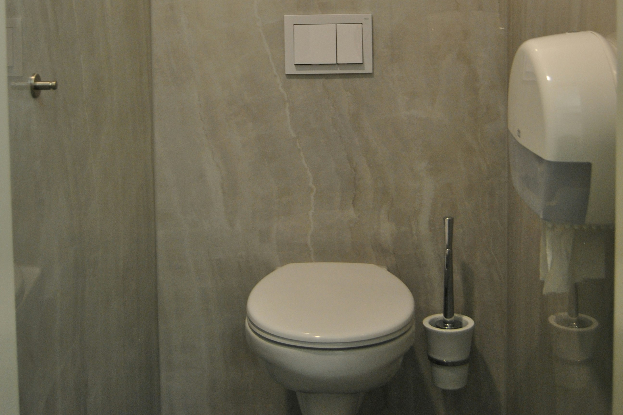 Weiße Toilette im modernen Badezimmer mit beigem Fliesenboden, Spülknopf, Wandarmatur und Seifenspendern.