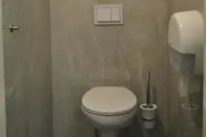 Weiße Toilette im modernen Badezimmer mit beigem Fliesenboden, Spülknopf, Wandarmatur und Seifenspendern.
