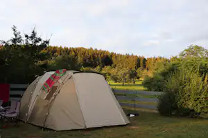 Zelt auf einem Campingplatz im Grünen, mit Stuhl und Blick auf Hecken, Wiese und einen Wald im Hintergrund.