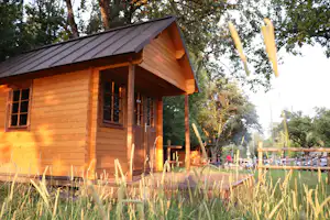 Holzhütte am Campingplatz mit Terrasse und Stühlen, umgeben von hohem Gras und Bäumen im Hintergrund.