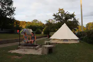 Campingzelt auf einer Stellfläche mit Sitzbank und Steintreppe, im Hintergrund Bäume und ein Weg.