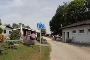 Campingplatz-Gehweg zwischen Gebäuden, links Rasen und Sitzbank, rechts Wohnwagen und geparkte Autos an der Straße.