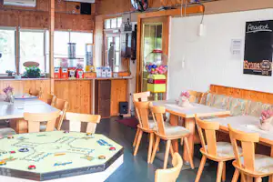 Spielzimmer mit Holztischen, Stühlen und einem Gumball-Automaten an der weißen Wand, daneben Schilder und Getränke-Boxen.