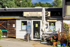 Kiosk & Café „Am Steinrodsee“ mit Rezeption am Campingplatz, Sonnenschirm, Blumenbeete und Schildern an der Fassade.