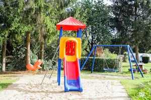 Bunte Rutsche und blaue Schaukeln auf einem Sandspielplatz im Grünen, mit Bäumen und Hecken im Hintergrund.