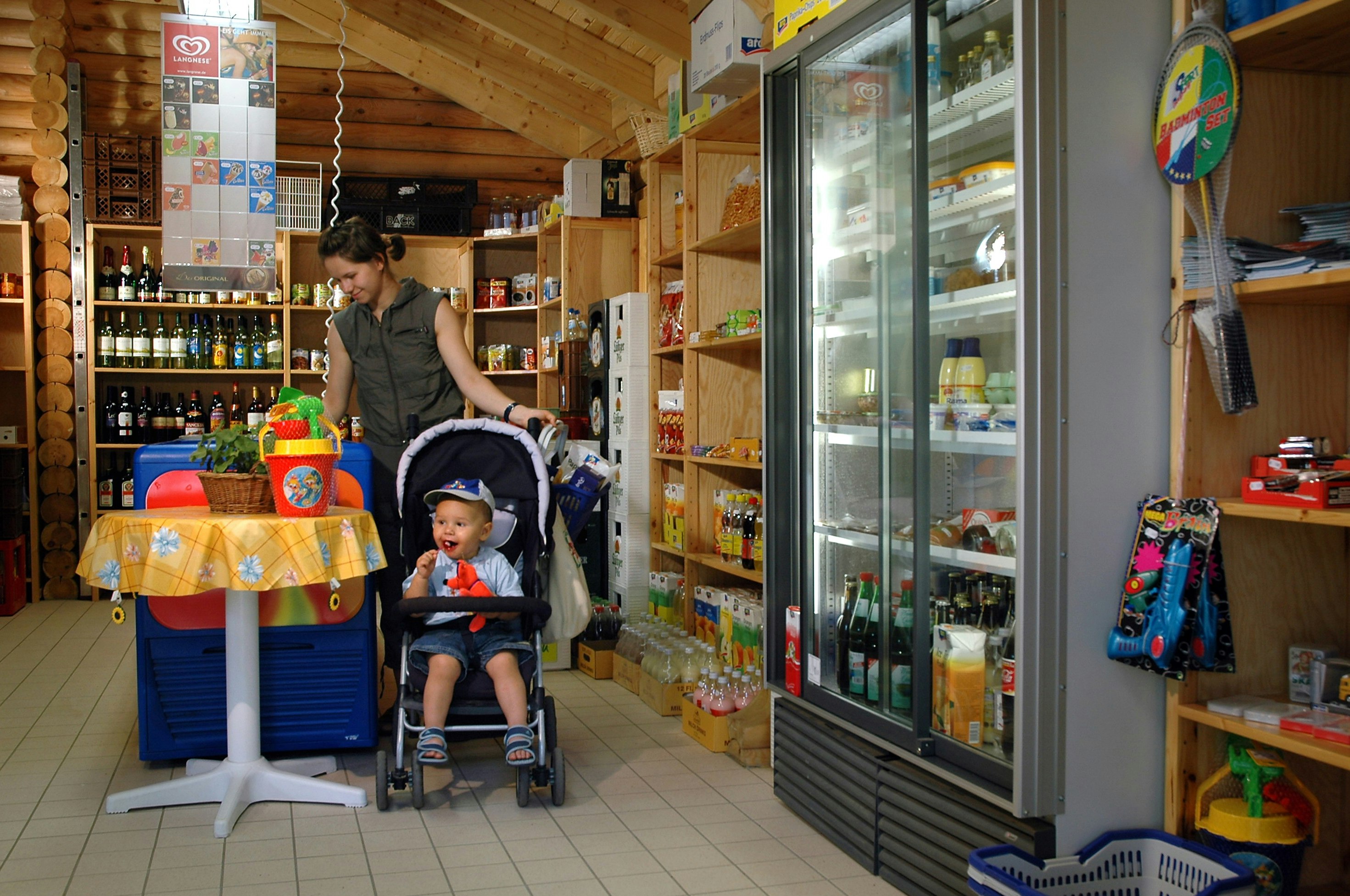 Camping-Shop in einem Holzgebäude: Regale mit Getränken, Kühlschrank, Tisch mit Decke und Kinderwagen im Vordergrund.