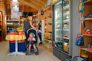 Camping-Shop in einem Holzgebäude: Regale mit Getränken, Kühlschrank, Tisch mit Decke und Kinderwagen im Vordergrund.