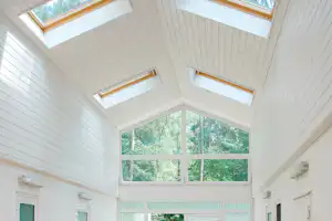 Flur in einem Campinghaus mit großen Fenstern zum Wald, hellen Wänden und Bodenfliesen, mehrere Personen gehen hindurch.
