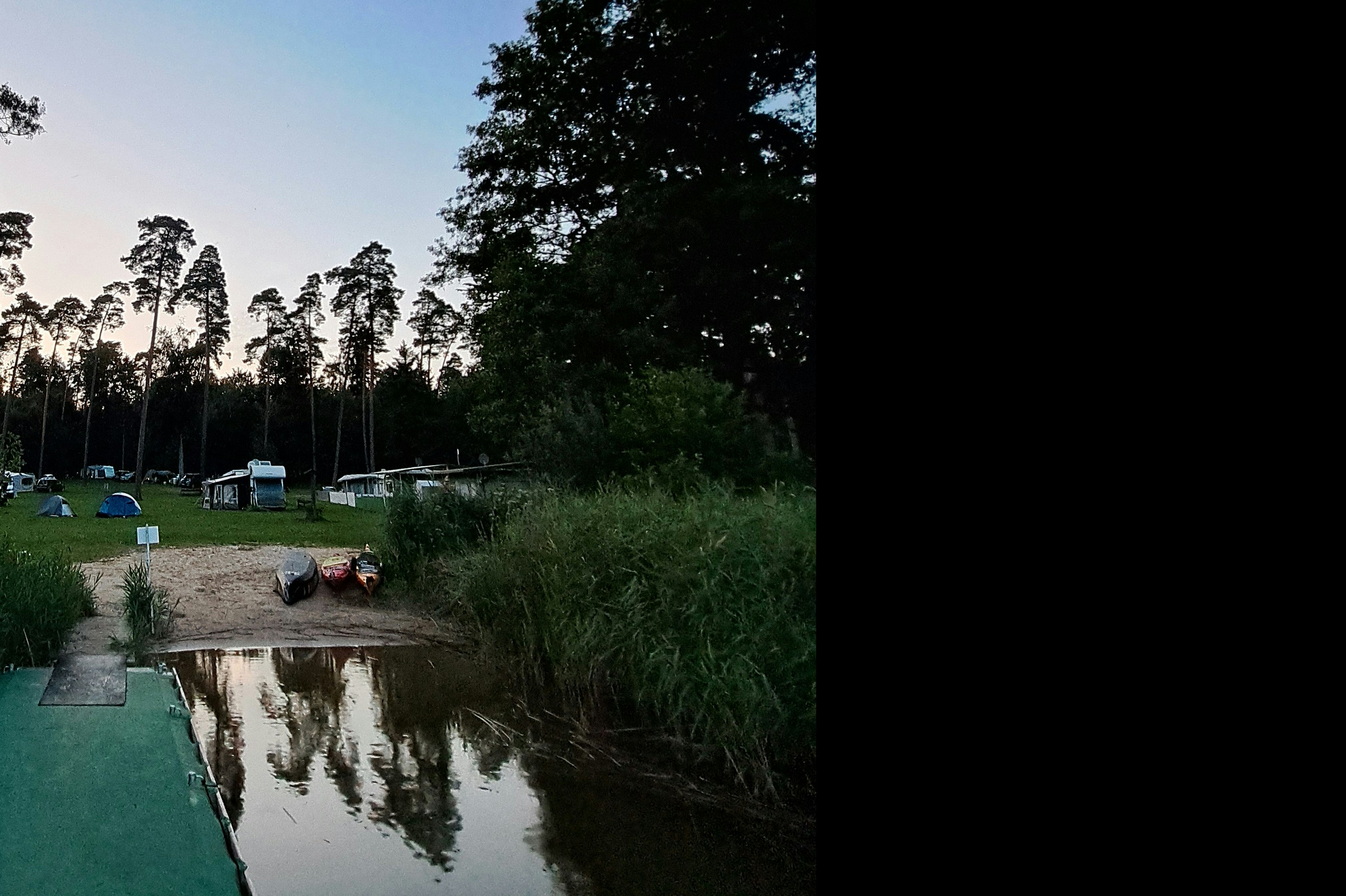 Campingplatz mit schmalem Weg am Wasser, Zelte und Wohnwagen im Hintergrund, Bäume und Gras entlang des Ufers.