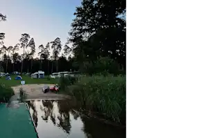 Campingplatz mit schmalem Weg am Wasser, Zelte und Wohnwagen im Hintergrund, Bäume und Gras entlang des Ufers.