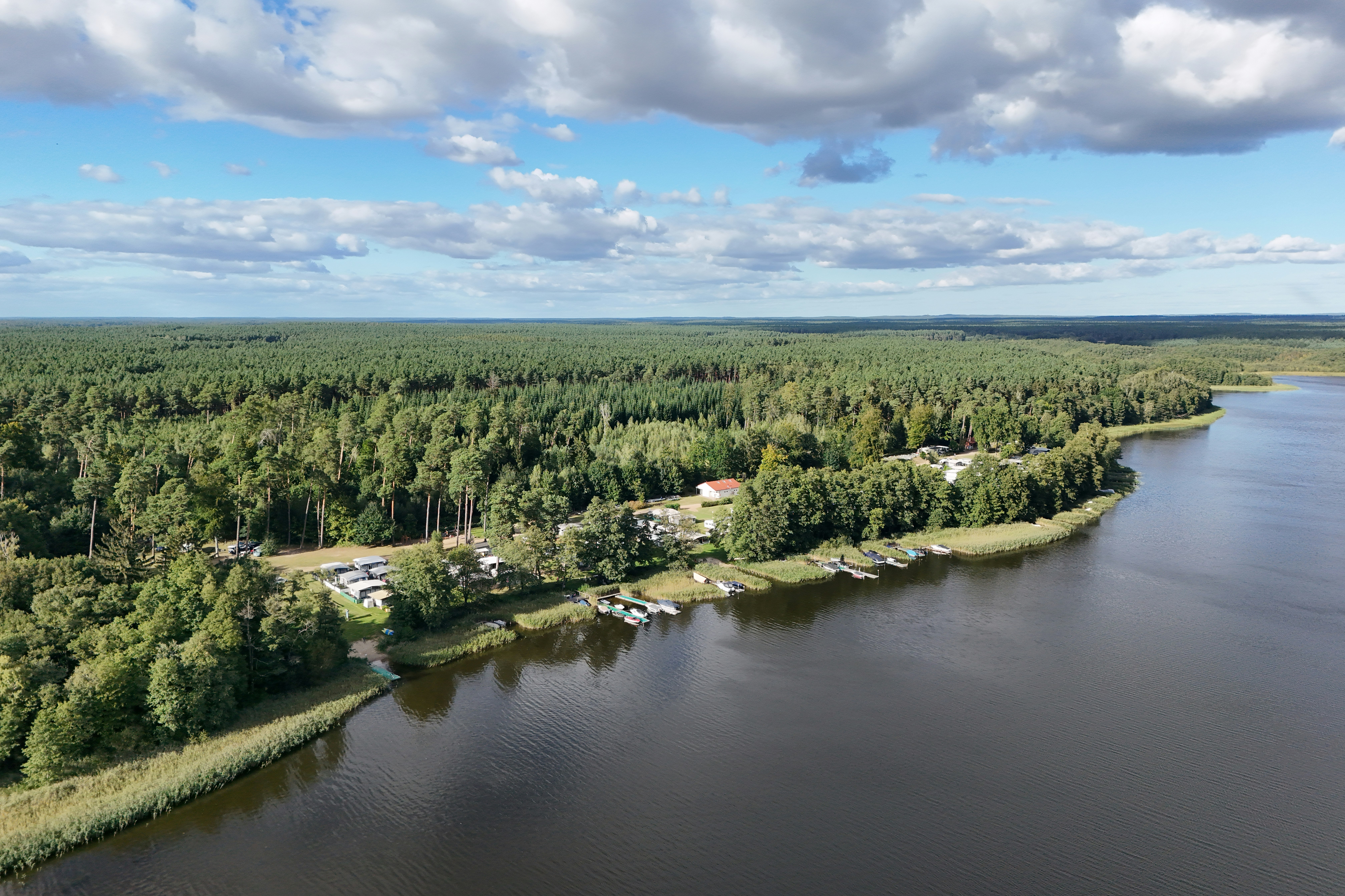 Luftaufnahme eines Flusses mit Uferwegen, Bootsstegen und Campingplätzen im Grünen, umgeben von Wald und Wolken am Himmel.