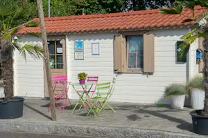 Wohnwagen mit Terrasse, bunten Klappstühlen und Tisch, Fensterläden und weißen Fassadenpaneelen, umgeben von Palmen und Kübeln.