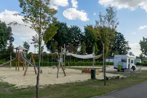 Spielplatz mit Holzgerüst und Seilnetz, daneben Wohnwagen und Wohnmobil auf dem Campingplatz, umgeben von Bäumen.
