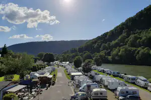 Campingplatz mit Wohnmobilen und Stellplätzen entlang eines breiten Weges, im Hintergrund bewaldete Hügel und ein Seeufer.