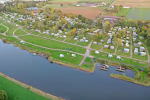 Luftaufnahme eines Campingplatzes am Fluss mit Stellplätzen, Wohnwagen und Wegen entlang des Ufers.