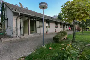 Reihenhaus am Campingplatz mit Terrasse, großen Fenstern, Pflanzenkübeln und gepflastertem Weg im grünen Garten.