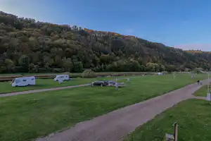 Wohnmobile und Autos auf einem Campingplatz auf grüner Wiese, mit Weg und Waldhang im Hintergrund.