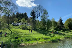 Grüne Wiese am Fluss mit Wohnmobilen und kleinen Gebäuden im Hintergrund, umgeben von Bäumen und Wald.