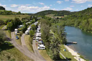 Campingplatz am Fluss mit Stellplätzen für Wohnmobile und Autos, Weg entlang des Ufers und Steg am Wasser.