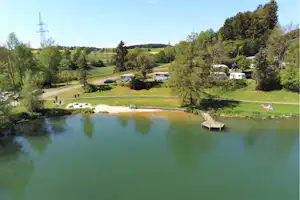 Campingplatz am See mit Rasenflächen, Stellplätzen und Wohnwagen, im Hintergrund Bäume und Hügel.