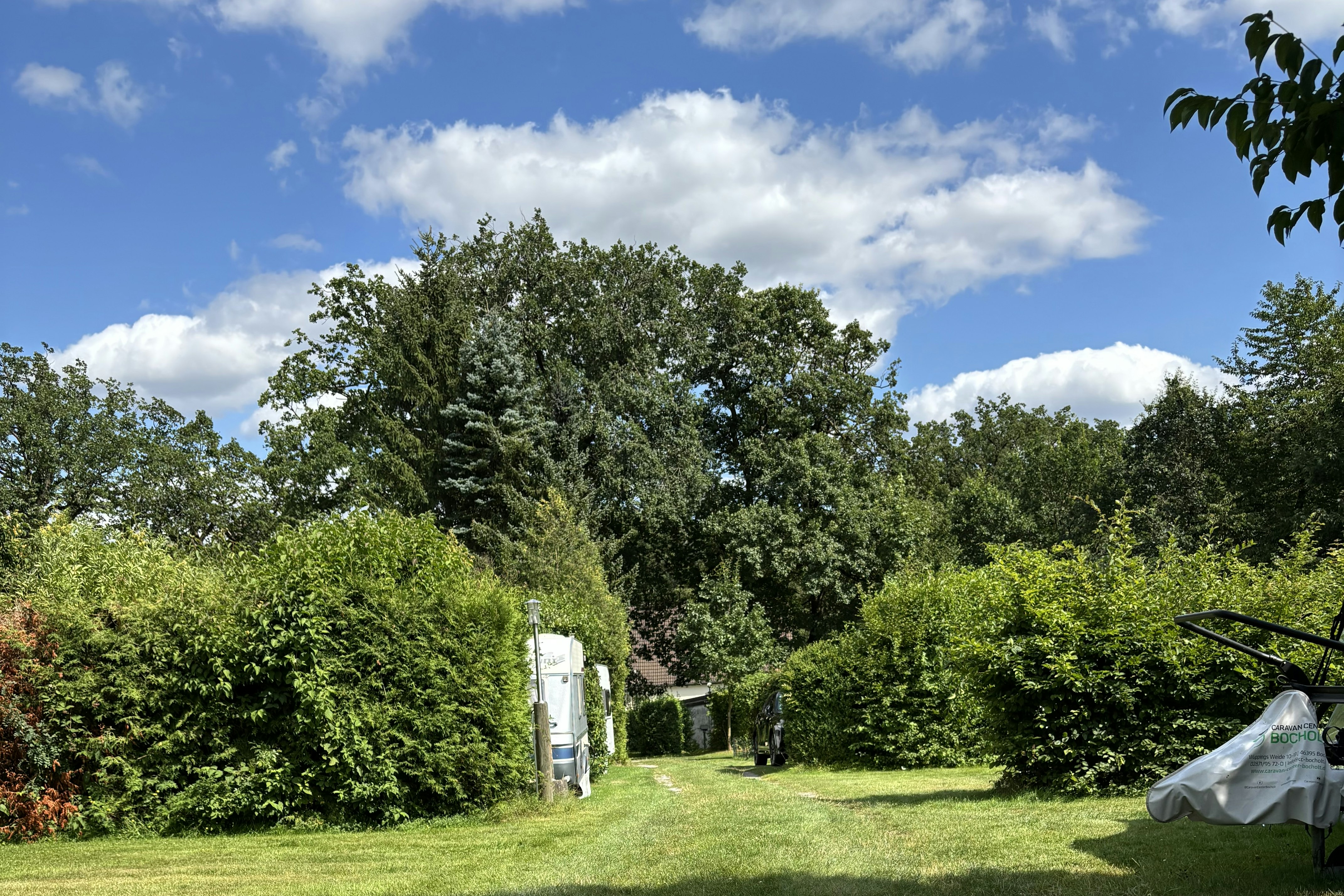 Grüner Campingplatz mit Rasenfläche, Hecken und Bäumen; rechts ein Zelt, im Hintergrund ein Wohnwagen und ein Weg.