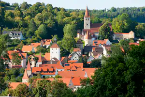 Kirche mit rotem Dach und Türmen über einem Dorf mit roten Ziegeldächern, umgeben von grünen Bäumen und Hügeln.