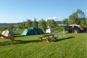 Campingwiese mit mehreren Zelten, einer Holzliege und bunten Markisen, im Hintergrund Bäume und Hügel.