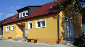 Gelbes Campinggebäude mit rotem Dach, Eingangstür und Fenstern, davor Bank und Weg auf dem Parkplatz.
