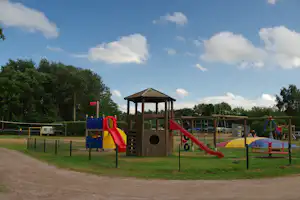 Spielplatz am Campingplatz mit Rutsche, Klettergerüst und Sandfläche, im Hintergrund Bäume und ein Wohnwagen.