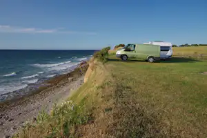 Grünes Wohnmobil steht am Strand auf einer Wiese, mit Blick auf das Meer und die Küste.