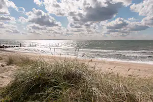 Sandstrand mit Dünen und Strandgras, dahinter das Meer mit Wellen und ein bewölkter Himmel.