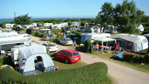 Campingplatz mit Wohnwagen und Zelten, Autos auf dem Stellplatz, Hecken und Bäume, im Hintergrund das Meer.