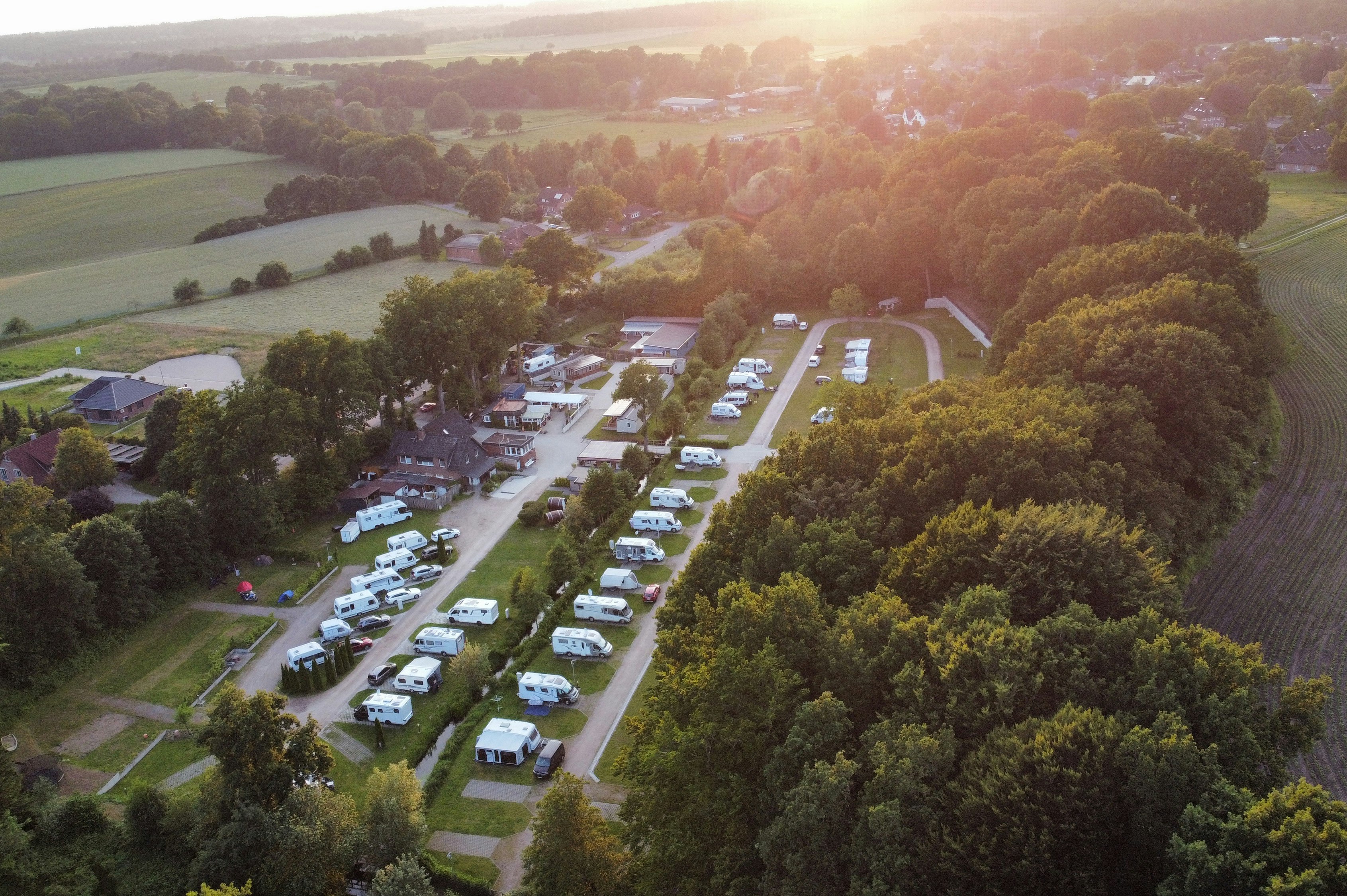 Luftaufnahme eines Campingplatzes mit Wohnmobilen und Stellplätzen entlang eines Weges, umgeben von Feldern und Bäumen.
