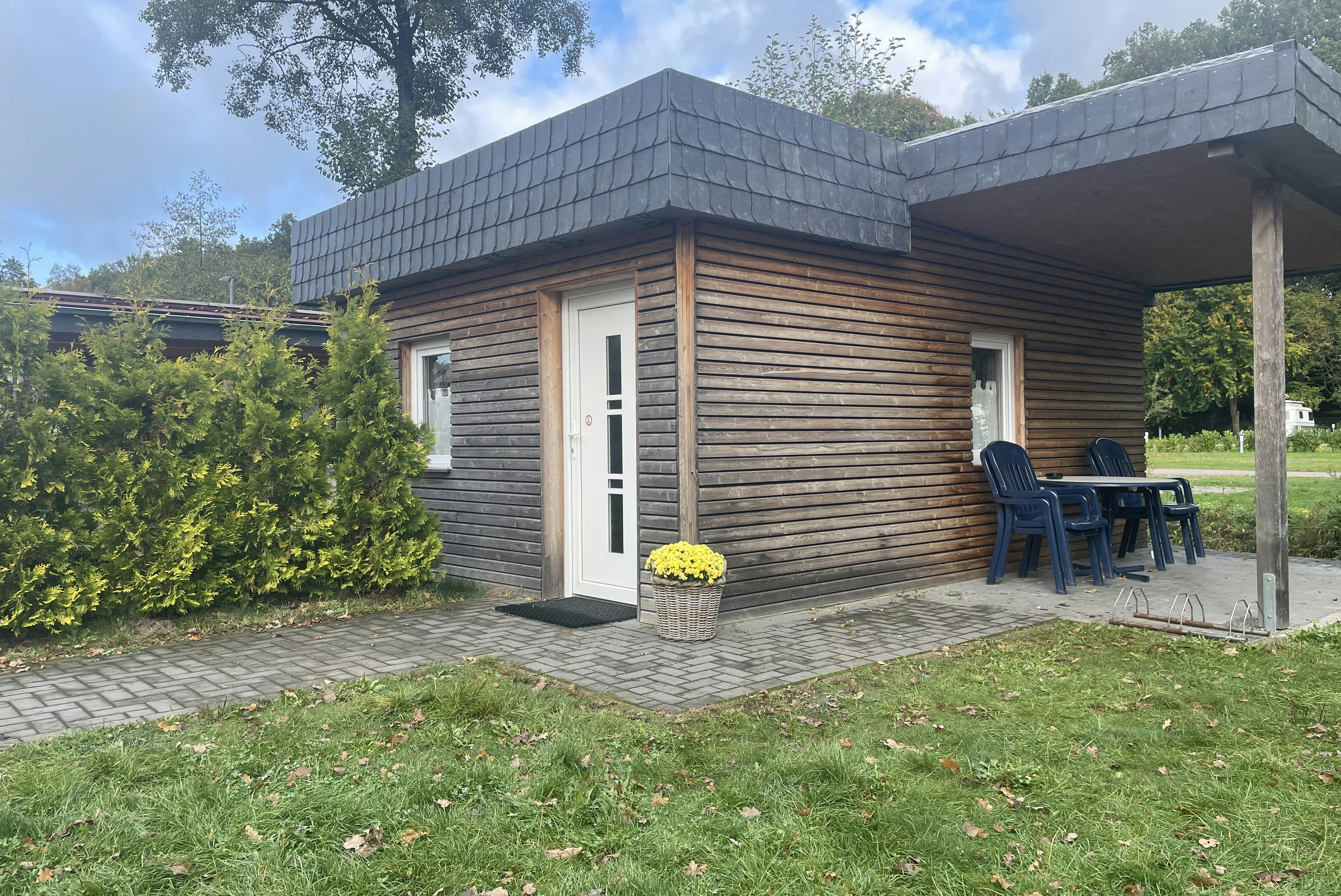 Holzbungalow mit Terrasse, zwei Stühlen und Tisch, gepflasterter Weg und Rasen im Garten, umgeben von Bäumen und Sträuchern.