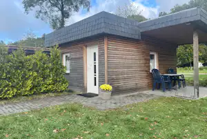 Holzbungalow mit Terrasse, zwei Stühlen und Tisch, gepflasterter Weg und Rasen im Garten, umgeben von Bäumen und Sträuchern.