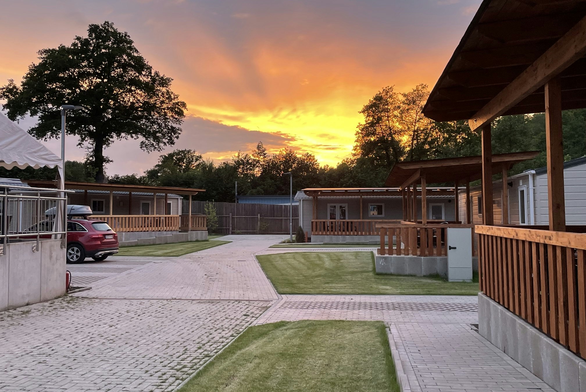 Campingplatz mit gepflastertem Weg, Rasenfläche und Holzunterstand am Rand, im Hintergrund Wohnmobile und Zaun bei Sonnenuntergang.